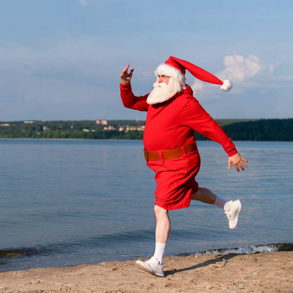 Santa Claus In Shorts Runs Along The Beach. Concept Of Celebrati Santa Claus in shorts runs along the beach. Concept of celebrating christmas at sea